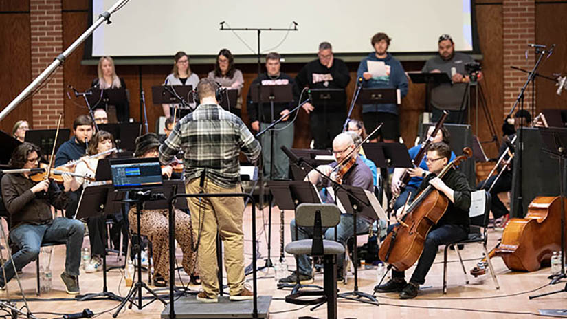 Photo of UW-River Falls student composer Sam Nesbit, at the podium, guiding a concert band of student and community musicians during a daylong cinema scoring event in Abbott Concert Hall. Nesbit and fellow students Ben Garcia and Landon Zientara wrote original music compositions to some of their favorite videogames and the gathered musicians performed them in time with key sections of each game. UWRF photo.